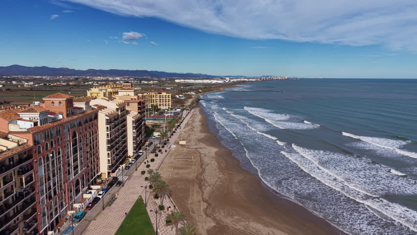 Aerial panoramic view of Port Saplaya in Alboraya, Valencia, showing beachfront apartments, palm-lined promenade, and rolling Mediterranean waves along the Spanish coast