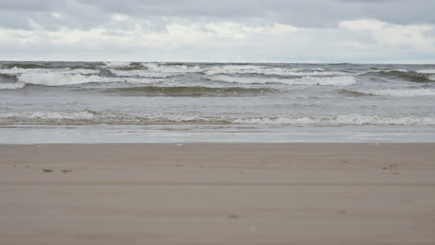 Sea waves rolling on sandy beach shore under overcast gray sky. Baltic sea coastline landscape with water motion and nature background. Cold windy weather at the seaside in autumn season.
