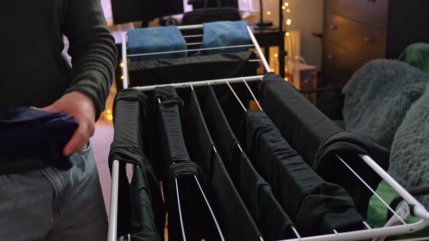 Man hanging navy briefs on an indoor drying rack, surrounded by dark clothes in a cozy apartment lit by warm fairy lights.
