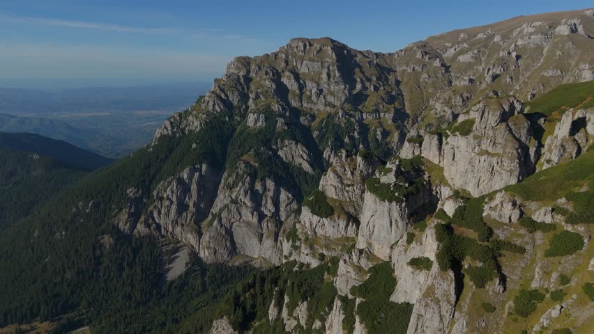 4K aerial drone footage continuing a slow, majestic flight over the deep Gaura Valley in the Bucegi Mountains, Romania. The camera gimbal stabilizes a smooth forward motion, showcasing the vast scale of the glacial cirque bordered by the rugged slopes of Gutanu and Lancia. The scene captures the interplay of light and shadow on the erosion towers and the patches of Dwarf Mountain Pine (Pinus mugo) and Alpine pasture below.