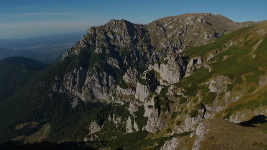 Spectacular 4K aerial drone footage performing a distant panoramic lateral reveal of the Gaura Valley in the Bucegi Mountains, Romania. Starting from the rugged Gutanu Mountain, the camera gimbal tracks towards the valley center, showcasing the imposing silhouettes of Lancia and Scara mountains. The scene captures numerous limestone erosion towers illuminated by the warm, soft light of the golden hour, creating a dramatic alpine atmosphere over the Dwarf Mountain Pine (Pinus mugo) covered slopes.