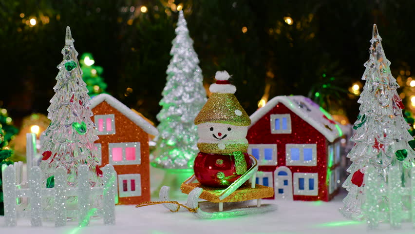 Two houses and a snowman on a sled and fir trees behind a fence against the backdrop of large fir branches