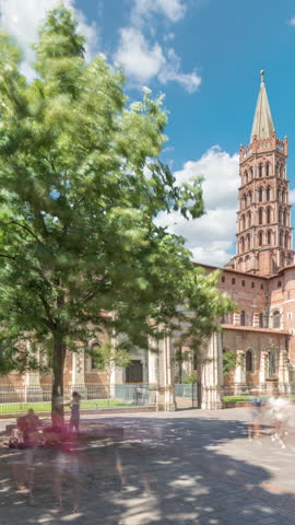 The cobbled parvis of the Basilica of Saint Sernin in Toulouse, France, timelapse hyperlapse. Largest romanesque church in Europe with brick bell tower, arched doors, green trees and blue cloudy sky