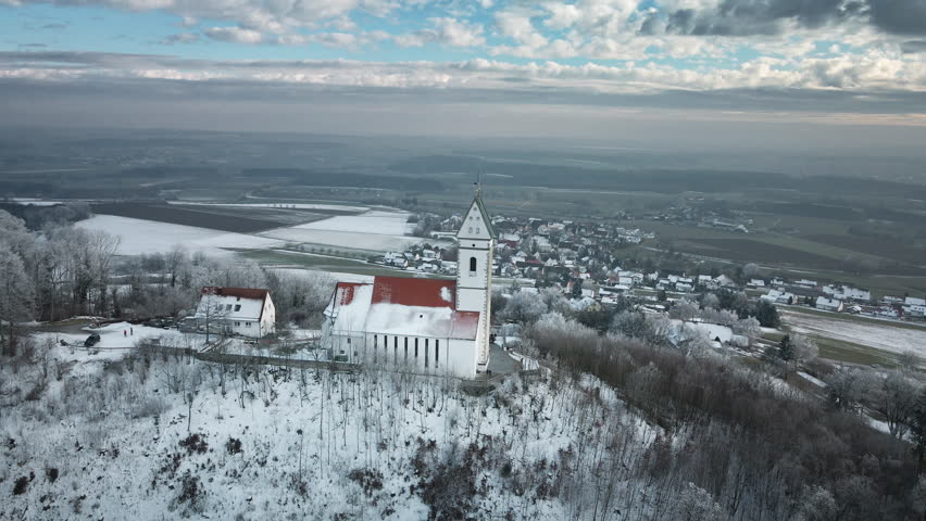 Drone initiates a second orbit circling the snow-covered white church with red roof and surrounding buildings on the hilltop in Utterweiler, Baden-Württemberg, amidst frosty trees