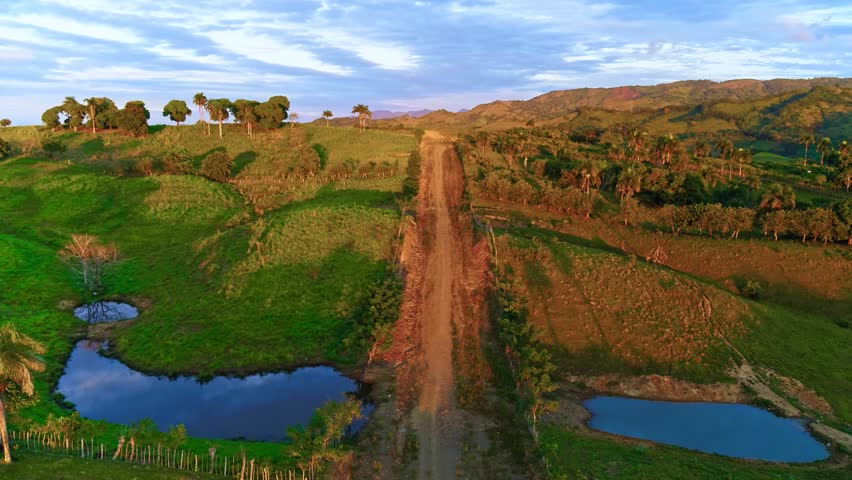 A view of a gravel country road in a mountain range with dense tropical forest and lakes. A Dominican Republic wilderness landscape at sunset. A landscape of mountain hills in the evening sunlight.