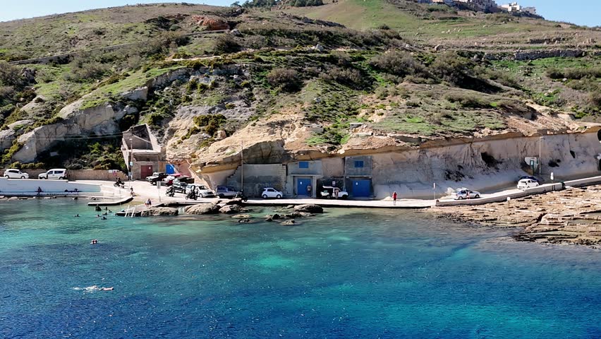 afternoon at Daħlet Qorrot Bay Gozo, Malta, featuring rustic limestone boathouses with vibrant blue doors carved into rugged cliffs overlooking a crystal-clear, shimmering turquoise Mediterranean Sea