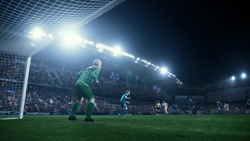 Soccer Player Hits the Ball and Scores a Goal. Goalkeeper Jumps in Opposite Direction. Stadium at Night with Bright Floodlights. Cinematic Slow Motion Footage from Inside the Gate