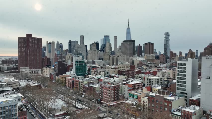 Aerial wide shot of New York City showing dense residential housing blocks and skyline with One World Trade Center in winter season under grey sky. Soft snow flurries falling from sky.