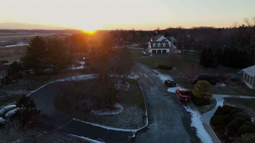 Quiet American suburban neighborhood at winter sunset, with frosty roads, parked cars and soft golden light illuminating homes and trees on cold evening in USA. Aerial approaching shot.