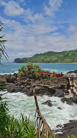 Scenic tropical coastline with rocky cliffs and lush green hills overlooking the blue ocean. Waves crash against the rugged shoreline under a bright sky with soft clouds, creating a refreshing and peaceful coastal atmosphere. This natural seaside landscape is ideal for travel, nature, environmental themes, and scenic background visuals.