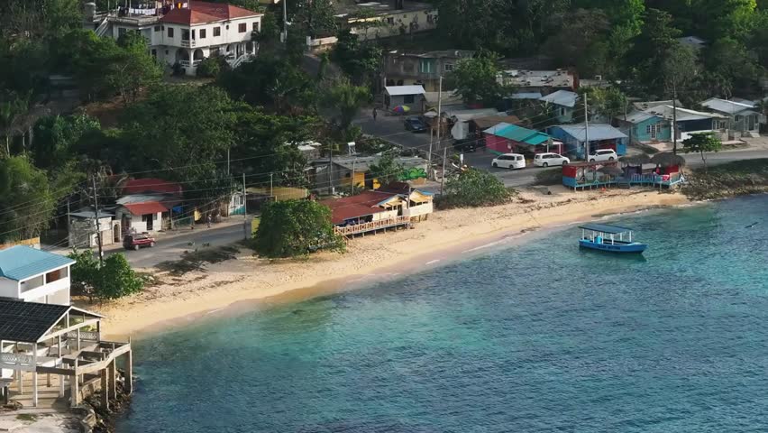 Aerial view of a tranquil Jamaican beach village, featuring turquoise waters, colorful homes, and a boat along the scenic coastline.