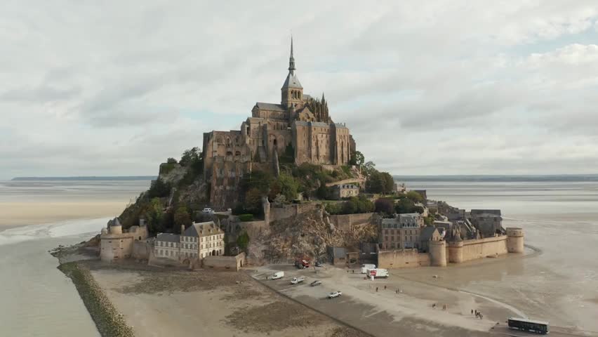 Wide aerial view of Le Mont Saint-Michel in France, rising from the ocean under cloudy skies, showcasing the historic castle and tidal island.