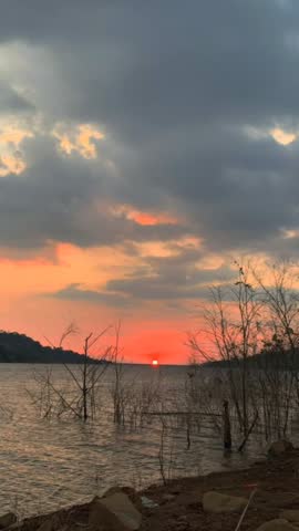 This image captures a dramatic sunset on the shore of a lake or reservoir. The sun appears as a bright orange ball, almost sinking into the horizon, creating a contrasting red and orange hue against the gray clouds above. In the foreground, dry tree branches jut out from the still water, creating a sense of stillness, poetry, and peace amidst the natural beauty