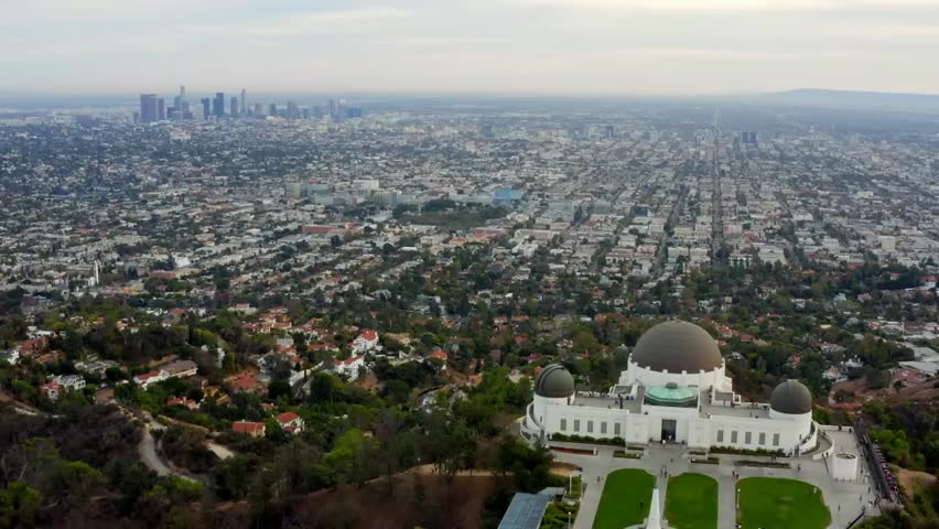 Aerial view over Griffith Observatory with Los Angeles skyline in the background, capturing the iconic landmark and cityscape in daylight under clouds.