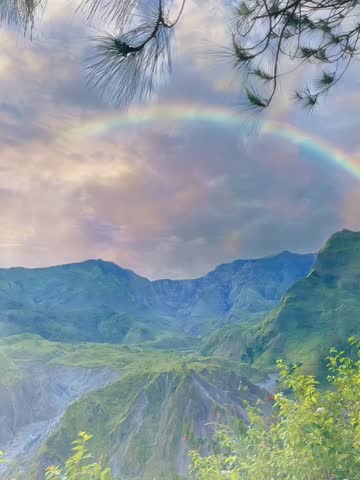 This image showcases a stunning mountain landscape, with a double rainbow arching beautifully over a green valley. The sky is dotted with soft pastel clouds at dusk or dawn, creating a magical and serene atmosphere. In the foreground, pine tree branches provide a natural frame to the composition, while the hills below reveal the dramatic contours of the land, blending lush green with rocky terrain. The overall image exudes spectacular natural beauty and serenity.
