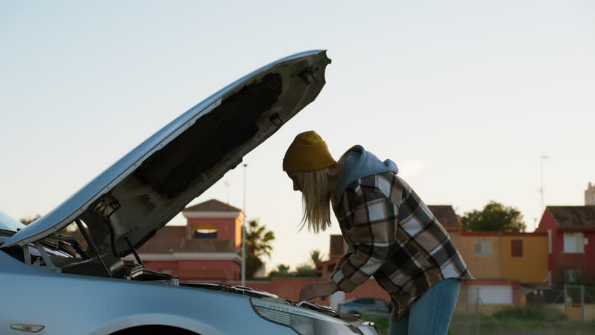 Young woman stranded at roadside, inspecting car open hood with worried expression while seeking help for engine problem and awaiting roadside assistance. Road problem, car trouble concept.