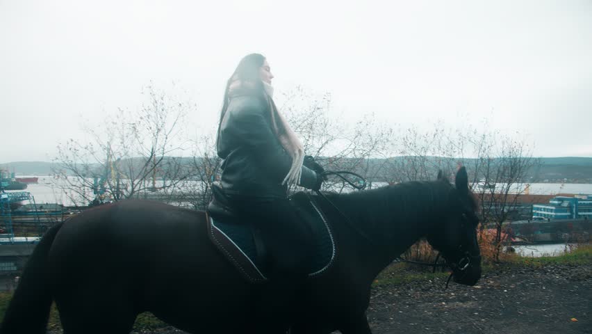 A long shot of an adult woman wearing a jacket and scarf, sitting astride a black horse, holding the reins. They walk against the backdrop of an autumn landscape and an overcast sky in countryside