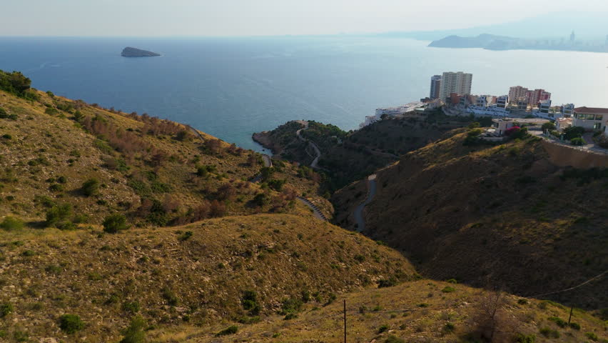 Scenic coastal landscape featuring rolling hills, distant island, and urban development, captured with a smooth dolly camera movement showcasing the serene seascape