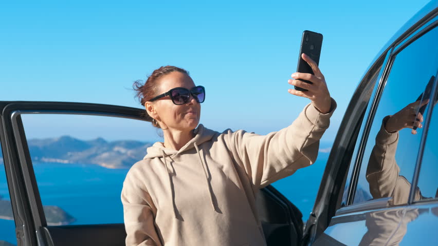 Woman taking selfie photo with phone during road trip. Happy woman taking a selfie with her smartphone next to her car on a mountain road with a stunning sea view in the background, enjoying her trip