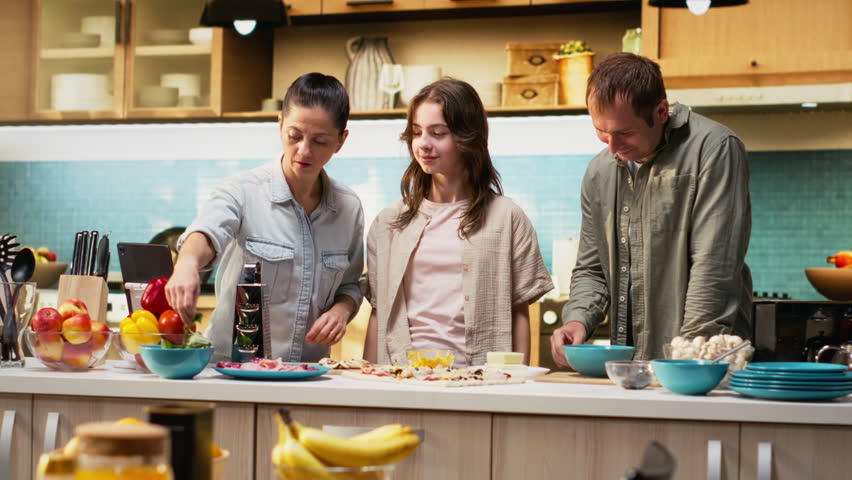 Mother takes selfies on phone while they cook delicious homemade pizza together in kitchen, daughter enjoying laughter and the warmth of family bonding with her parents. Camera B.