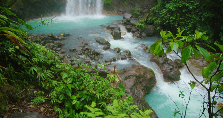 Tourist woman exploring Costa Rica waterfall pool surrounded by bright turquoise river in tropical rainforest during the rain