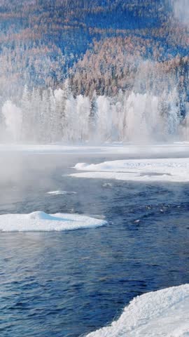 Vertical 1080p video of a flowing unfrozen river in a snowy winter landscape. Steam and mist rise from the cold water, creating a dreamlike atmosphere. The riverbanks are lined with trees covered in white rime ice (frost), while the background mountains feature brownish larch forests. Scenic nature footage, likely resembling Kanas in Xinjiang or Northern China.