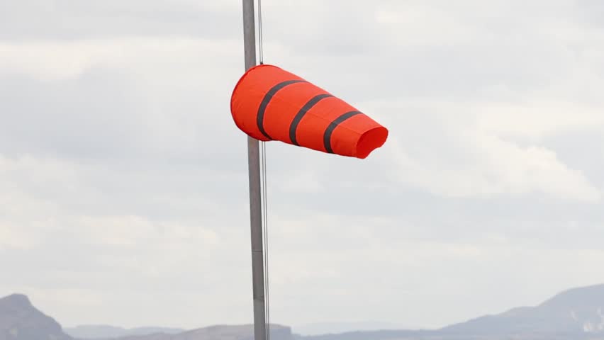 A vibrant orange windsock sways on a pole against a backdrop of hills and clouds.