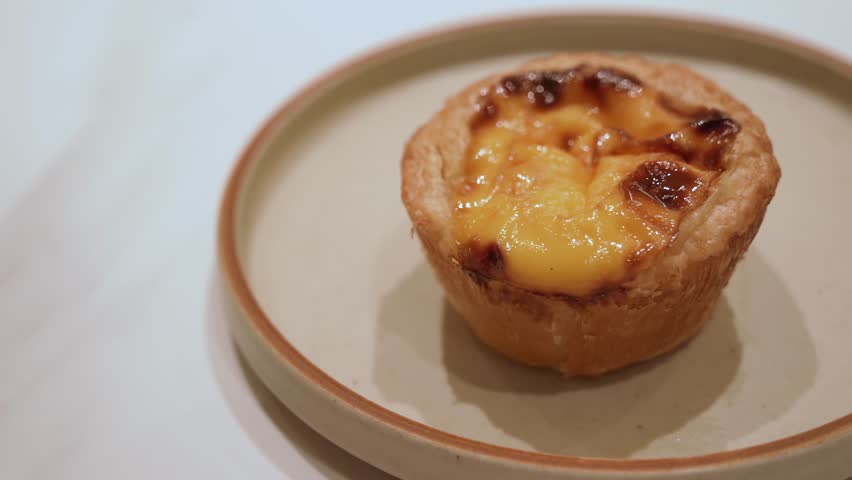 A small egg tart pastry served on a neutral plate, featuring a golden, caramelized custard filling.