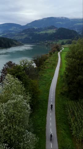 Couple rides ebikes along Drava River bike path through green fields, vertical drone follow shot