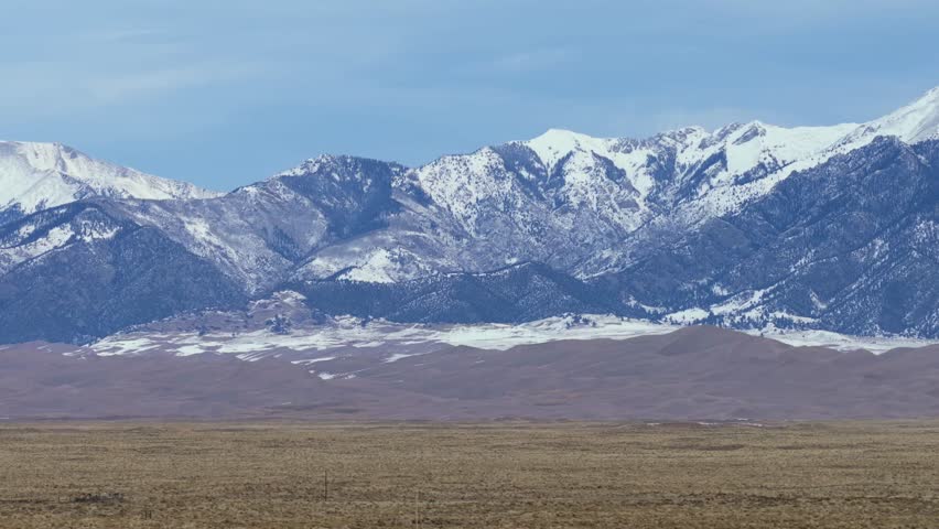 High-altitude winter sunlight illuminates the Sangre de Cristo Mountains and the Great Sand Dunes of Colorado. This wide aerial panorama captures the epic scale of the snow-covered peaks and desert.