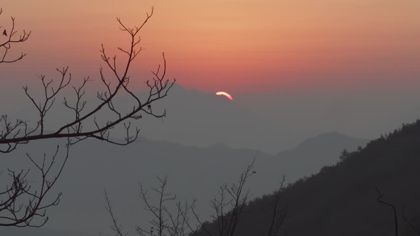 4K fast motion footage of sunrise viewed from top of mountain, sun rising behind mountains far away, golden sun with orange sky background and silhouette of tree branch foreground, peaceful landscape