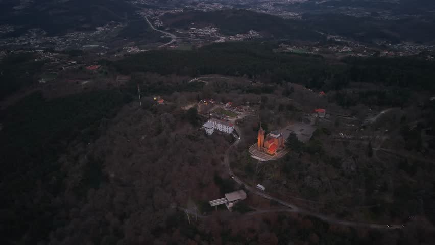 Aerial sunset view of Penha sanctuary surrounded by forested hills, warm golden light, winding roads and city of Guimarães in the background.