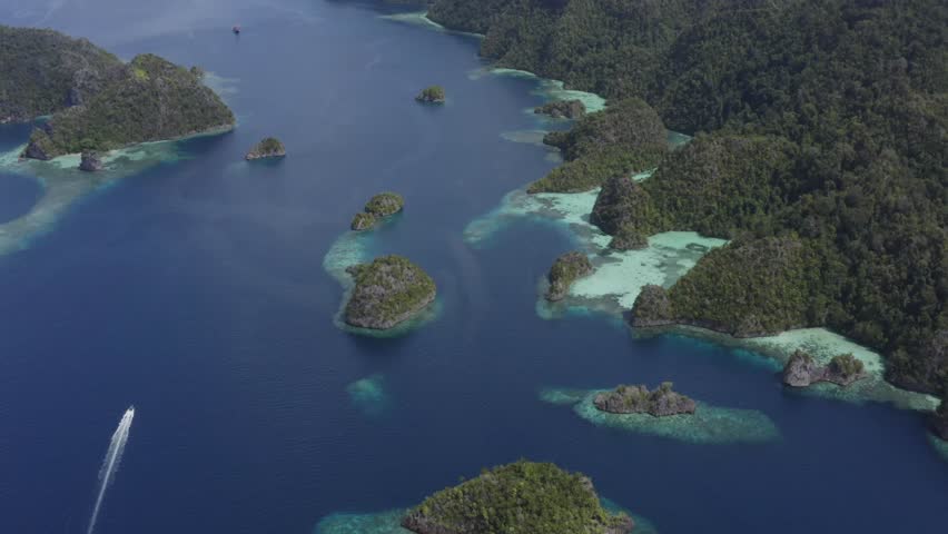 Aerial view of a  speed boat cruising through a channel between karst islands surrounded by blue water and coral reefs, Misool, Raja Ampat, Indonesia, Pacific Ocean
