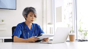 Healthcare worker in scrubs sits at desk, holding a  patient medical charts , rubbing her temple and showing fatigue, stress and burnout from medical frontline pressures - Powered by Shutterstock - Get 15% off with code: PIKWIZARD15