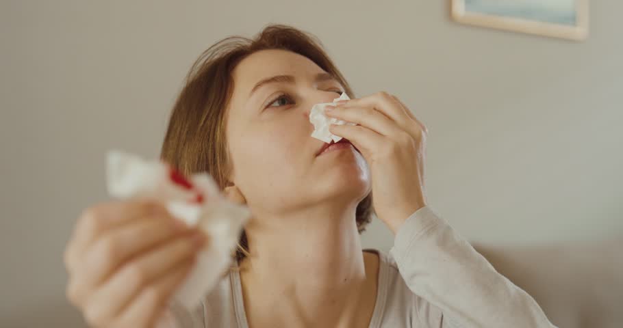 A woman sitting at home treating a sudden nosebleed with tissues — concept of minor health issue, first aid, and everyday medical care.