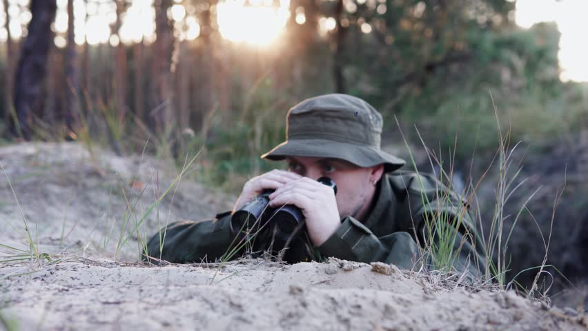 Soldier in a trench scans enemy lines with binoculars during a training exercise.