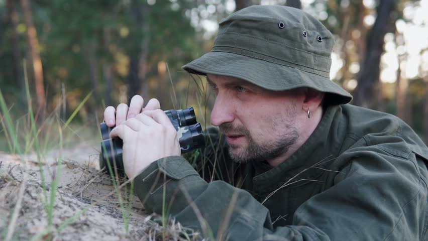 Soldier in a trench scans enemy lines with binoculars during a training exercise.