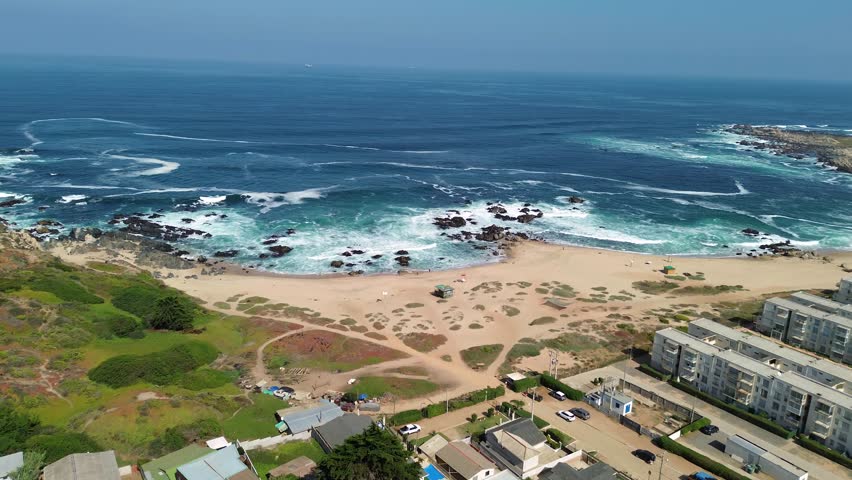Drone zoom-in shot over the coastal town of El Tabo, located on the central coast of Chile in the Valparaíso Region. Ocean waves, sandy beach, rocky shoreline and residential houses by the sea