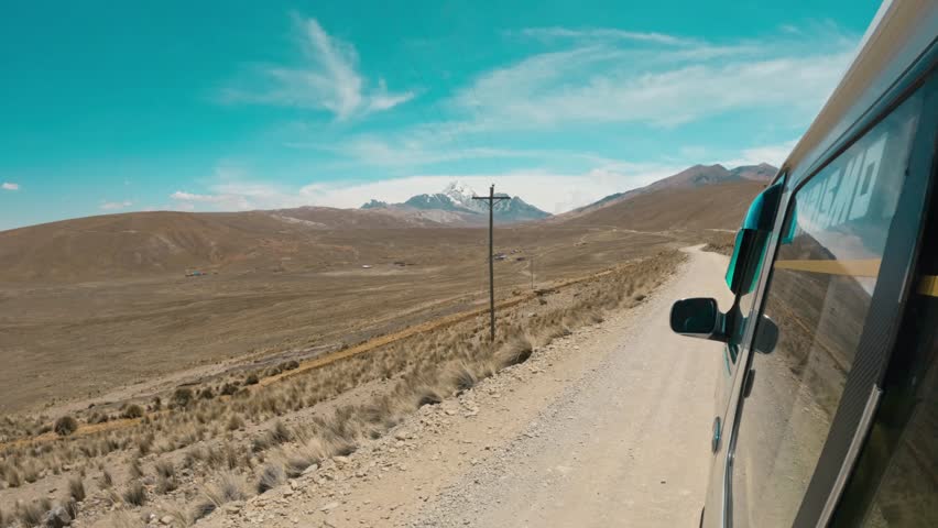 View from a tourism van driving on a remote dirt road in the Bolivian Andes. The vast, arid Altiplano landscape leading toward high-altitude snow peaks.
