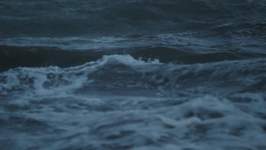Rough ocean waves rolling in slow motion after sunset with visible wind impact and pre storm atmosphere during blue hour.