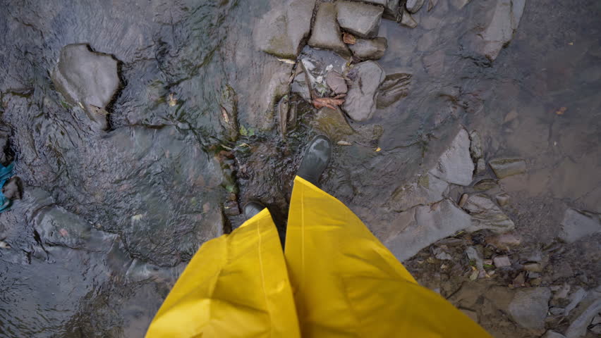 White woman pov stepping on stones, closeup boots and hem of raincoat, slippery rocks and shallow water, careful footing, field scientist checking flow, rugged