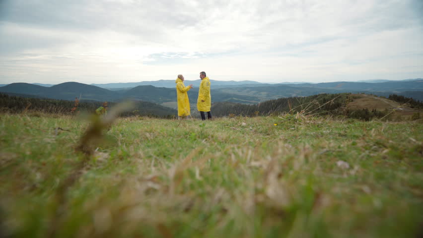 two people yellow raincoats standing grassy hill overlooking distant mountains and cloudy horizon, softfocus foreground grass, overcast sky, subtle breeze,