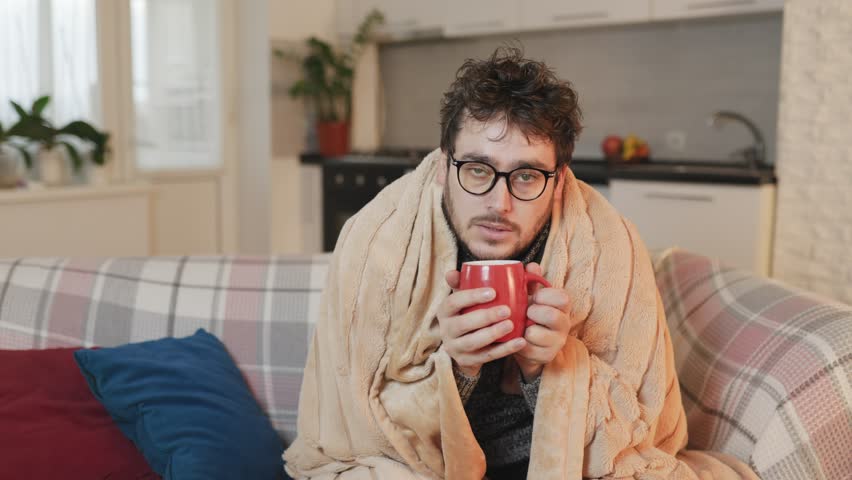 a young man battles the freezing temperatures at home in the living room. Donning eyeglasses and covered with a duvet, his sad demeanor reflects the harsh reality of cold,