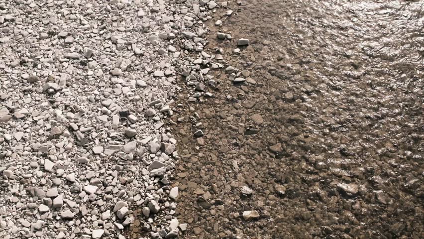 Shallow river water flowing along a rocky shoreline seen from above. Clear stream meets dry stones, forming a natural boundary with gentle motion and textured surface