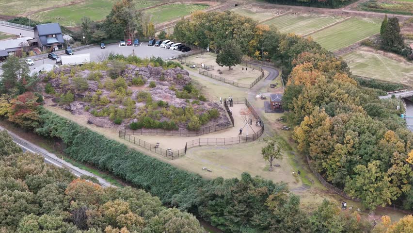 A park with a fence and a pond. Trees are in the background. There are people walking around the park