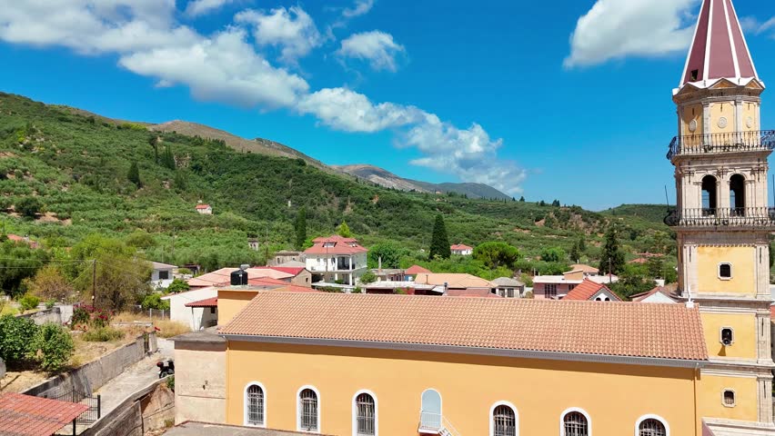 This aerial drone footage showcases the picturesque Kiliomenos Village in Zakynthos, Greece. The video features an overhead view of the Church of Agios Nikolaos, with its distinctive architecture and red roof, set against a stunning backdrop of lush greenery and rolling hills. The drone flies over the village, offering a unique perspective on the charming Greek landscape.