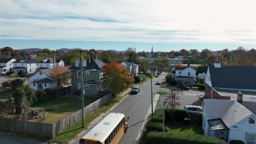 Aerial tracking shot of yellow school bus stopping at a quiet residential intersection in autumn, surrounded by colorful trees and traditional American houses in small town of USA.