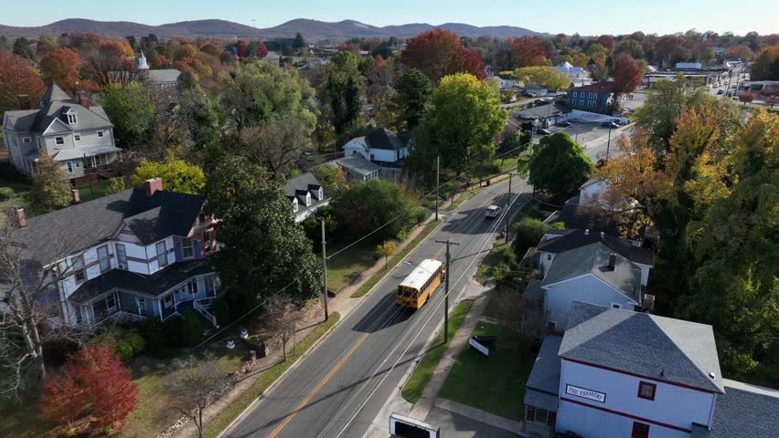 Aerial tracking shot of yellow school bus on street between colored trees in suburb housing area. Wide shot. Houses and homes in small town of USA.