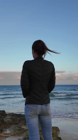 Woman with her back to the camera contemplating the waves of the sea from a rocky beach
