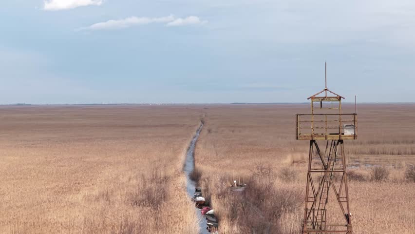 A vast wetland landscape covered with dry reeds stretches toward the horizon under a pale blue sky. A narrow water channel leads through the marsh, while a wooden watchtower stands on the right.